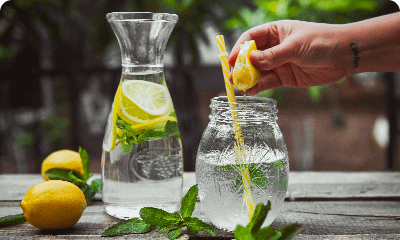 hand-squeezing-lemon-into-glass-jar-with-water-side-view-wooden-yard-table
